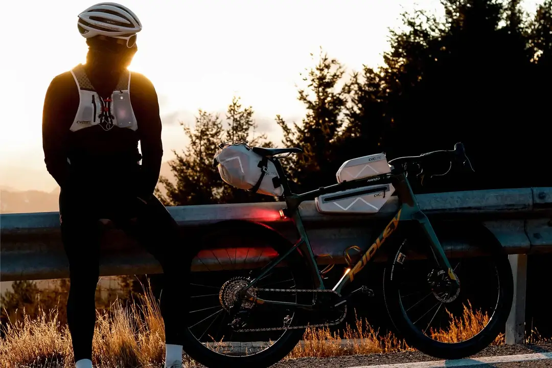 Image d'un homme en contre-jour assis sur une barrière avec à sa droite un vélo équipé de sacoche cyclite